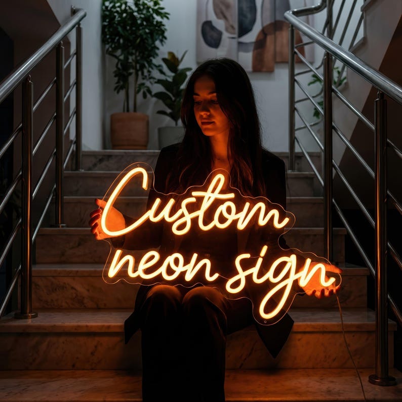 May include: A glowing neon sign with the words "Custom neon sign" in a cursive font. The sign is a warm orange color and is held by a person sitting on a staircase. The background includes a staircase, plants, and artwork.