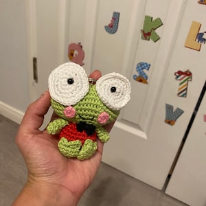 May include: A green crocheted frog keychain with white eyes, a red and black bow tie, and pink cheeks.