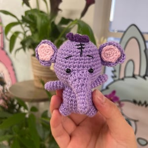 May include: A handmade crochet elephant in shades of purple, with pink inner ears and a small purple tuft on its head. The elephant has small black eyes and is held in a hand. The craftwork is detailed and the elephant is a cute, whimsical design.