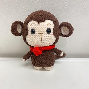 May include: A handmade crochet monkey doll with a brown body, head, and limbs. The monkey has a cream-colored face, ears, and feet, with black eyes and a red scarf. The doll is sitting on a white surface.