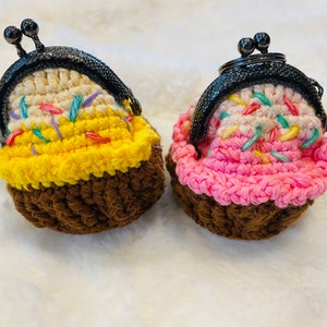 May include: Two crocheted coin purses shaped like cupcakes. One is yellow with brown frosting and the other is pink with brown frosting. Both have colorful sprinkles.
