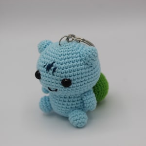 May include: A light blue crocheted plush toy with a green crocheted shell on its back. The toy has a key ring attached to its head and black embroidered eyes.