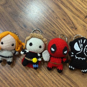 May include: Four crocheted character keychains. The characters are a blonde woman, a man with a black and red costume, a red and black character with a mask, and a black character with a white mask.
