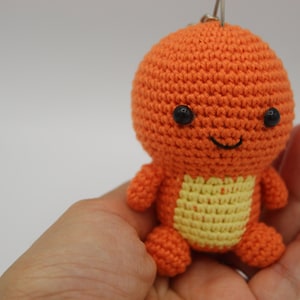 May include: Orange and yellow crocheted plush toy with a smiling face and black eyes. The toy is being held in a hand.