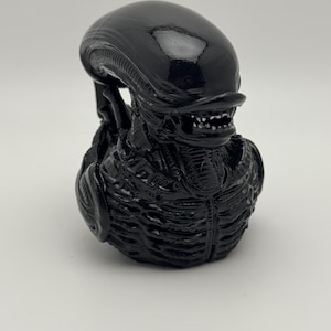 May include: A black alien bust figurine with a detailed, textured surface. The alien has a large head with a prominent jaw and sharp teeth.