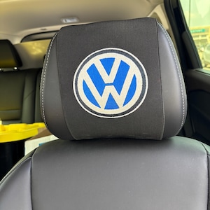 Könnte beinhalten: Ein schwarzer Kopfstützenbezug für das Auto mit einem kreisförmigen, in Blau und Weiß gestickten Volkswagen-Logo. Der Bezug ist über einer dunkelgrauen Lederkopfstütze im Fahrzeug angebracht und zeigt das VW-Emblem mittig auf der Vorderseite.