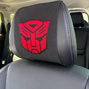 Peut inclure: Une housse d'appui-tête de voiture noire arborant un logo Autobot rouge brodé, issu de la franchise Transformers. La housse est fabriquée dans un tissu extensible et est ajustée sur la partie supérieure d'un appui-tête en cuir noir.
