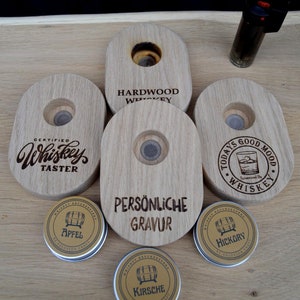 Può includere: Cinque tappi per bottiglia di whisky in legno con diverse incisioni. I tappi sono su una superficie di legno. Ci sono quattro piccole lattine rotonde di metallo con diverse etichette. Le etichette recitano "Whiskey Rauchspann" e mostrano un'immagine di una botte. Le etichette recitano anche "Apfel", "Hickory" e "Kirsche".