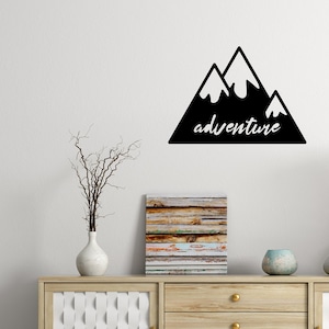 Könnte beinhalten: Schwarze und weiße Wanddekoration mit einer Bergsilhouette und dem Wort "Abenteuer" darunter. Die Dekoration ist an einer weißen Wand über einer Kommode aus Holz mit einer weißen und hölzernen Front angebracht.