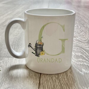 May include: White ceramic mug with a green letter G and the word "Grandad" printed on it. The letter G has a floral design and a watering can with a trowel.