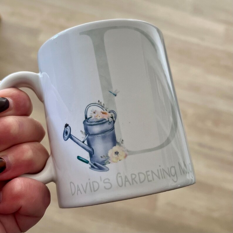 May include: White ceramic mug with a light blue watering can and flowers design. The mug has the text "David's Gardening Mug" on the bottom.
