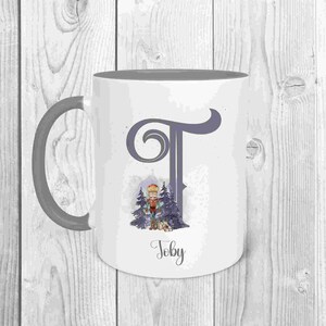 May include: A white ceramic mug with a grey handle. The mug features a grey and white illustration of a nutcracker in a winter scene with the letter "T" and the name "Toby" printed on the mug.