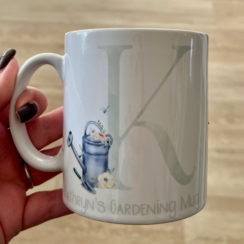 May include: White ceramic mug with a gray letter K and a watercolor illustration of a watering can with flowers. The mug says "Kathryn's Gardening Mug".