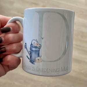May include: White ceramic mug with a green and white letter D, a blue watering can with white flowers, and the text "David's Gardening Mug".