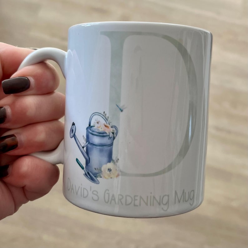 May include: White ceramic mug with a green and white letter D, a blue watering can with white flowers, and the text "David's Gardening Mug".