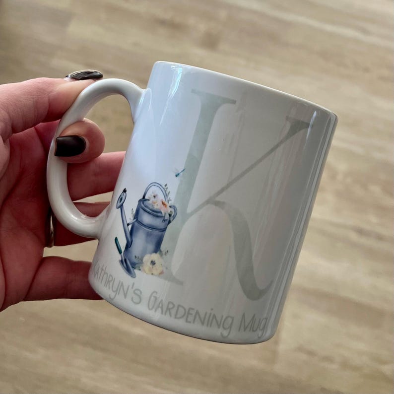 May include: White ceramic mug with a gray letter K and a watercolor illustration of a watering can and flowers. The mug has the text "Kathryn's Gardening Mug" on the bottom.