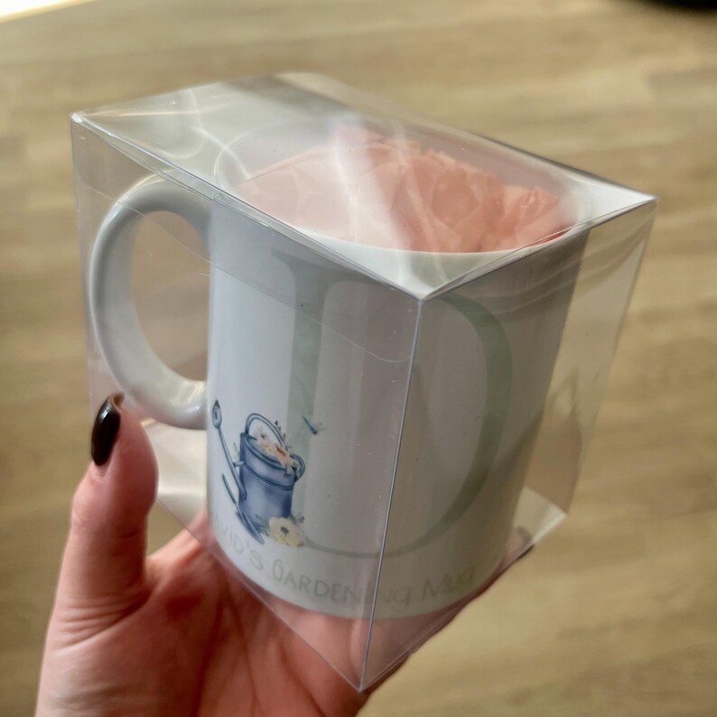 May include: White ceramic mug with a blue watering can and flowers design. The mug has the text "J's Garden" on the bottom. The mug is packaged in a clear plastic box.