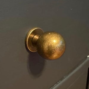 May include: A spherical cabinet knob with a metallic brass finish and a circular base plate. The knob features a smooth, rounded surface with a subtle weathered texture, mounted onto a dark, solid-colored flat surface that serves as a drawer front.