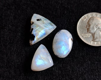 May include: Three iridescent moonstone cabochons in a triangular, oval, and triangular shape. The stones are a milky white with a blue sheen. The stones are next to a US quarter for size reference.