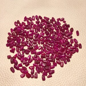 May include: A scattering of small, raw ruby gemstones. The gemstones are a deep, rich red colour and appear to be uncut. They are spread across a light-coloured surface, creating a visually appealing display of natural beauty.