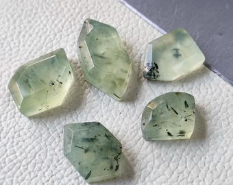 May include: Five faceted prehnite gemstones with a pale green hue and black inclusions. The gemstones are cut into various geometric shapes, showcasing their natural beauty. The stones are displayed on a white surface.