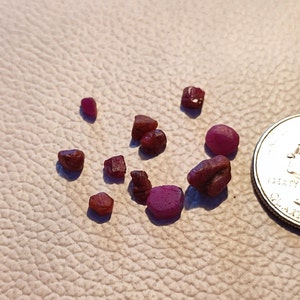 Puede incluir: Una colección de pequeñas piedras preciosas de rubí rojo, algunas con un tono ligeramente púrpura, esparcidas sobre una superficie blanca texturizada. Se incluye una moneda de veinticinco centavos de dólar estadounidense para la escala.