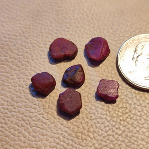 Puede incluir: Seis piedras preciosas de rubí rojo de varias formas y tamaños. Las piedras preciosas están dispuestas sobre una superficie texturizada de color marrón claro. Se incluye una moneda de veinticinco centavos de dólar estadounidense para la escala.