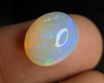 Può includere: Una gemma di opale ovale con una base bianca lattea. L'opale mostra lampi iridescenti di blu, verde e arancione. La gemma è liscia e lucida, riflettendo la luce.