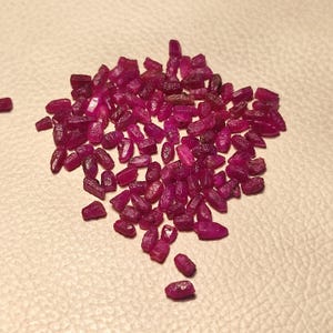 Könnte beinhalten: Eine Ansammlung kleiner, grob geschliffener Rubin-Edelsteine. Die Rubine haben eine tiefe, satte rote Farbe und wirken leicht durchscheinend. Sie liegen auf einer hellen Oberfläche verstreut.