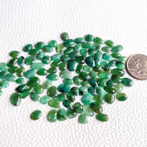 Könnte beinhalten: Eine Ansammlung von Smaragd-Edelsteinen in verschiedenen Formen, darunter oval und tropfenförmig. Die Edelsteine weisen eine Bandbreite an Grüntönen auf. Eine Münze dient als Größenvergleich. Maße in Zentimetern.