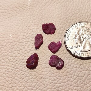Könnte beinhalten: Fünf kleine, raue, rosafarbene Rubingesteine auf einer hellbraunen Lederoberfläche. Eine US-Viertel-Dollar-Münze dient zum Größenvergleich.