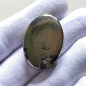 Può includere: Una pietra preziosa ovale liscia di colore grigio scuro con un bagliore metallico. La pietra ha una superficie leggermente strutturata ed è posizionata su uno sfondo bianco.