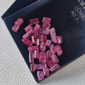 May include: A collection of small, pink, rectangular crystals. The crystals are scattered on a black surface, with some appearing translucent. The crystals have a natural, rough texture and varying sizes. The image is well-lit, highlighting the color and form of the crystals.
