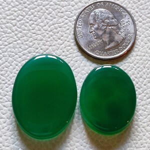 Può includere: Due cabochon di agata di forma ovale di colore verde, uno leggermente più grande dell'altro, su una superficie bianca testurizzata. Una moneta da un quarto di dollaro americano è inclusa come riferimento per le dimensioni.