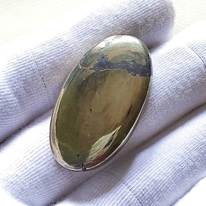 Può includere: Una pietra preziosa ovale liscia di colore oro metallico con una linea grigio scuro che attraversa il centro. La pietra preziosa è su uno sfondo di tessuto bianco.