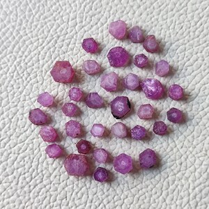 May include: A collection of small, rough-cut, pink gemstones scattered on a white surface. The gemstones vary in size and shape, with a range of light to dark pink hues. The overall arrangement is random, creating a natural and organic aesthetic.