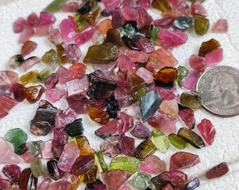 May include: A collection of small, colorful, and irregularly shaped gemstones. The gemstones are a mix of pink, green, brown, and black. The gemstones are scattered on a white surface.