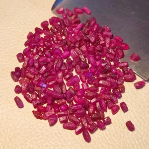 Op de afbeelding: Een stapel ruwe, ongeslepen robijn edelstenen in verschillende maten en vormen. De edelstenen hebben een diepe, rijke rode kleur, met een lichte paarse tint. Een metalen schep is gedeeltelijk zichtbaar in de rechterbovenhoek.