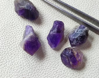 Peut inclure: Cinq cristaux d'améthyste bruts de différentes formes et tailles, affichant des nuances de violet et de pourpre. Une paire de pinces argentées est posée près des cristaux sur une surface blanche.