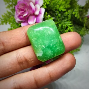 May include: A smooth, green, rectangular cabochon gemstone with a light green, translucent appearance. The stone has a slightly cloudy, marbled pattern.