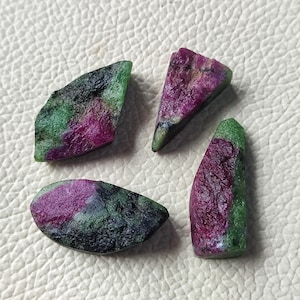 Könnte beinhalten: Vier rohe Rubin- und Zoisit-Edelsteine. Die Steine zeigen eine Mischung aus Farben, darunter tiefes Rot, Grün und Schwarz. Die Formen variieren, wobei einige Stücke dreieckig oder unregelmäßig erscheinen. Die Edelsteine haben eine raue, natürliche Textur.