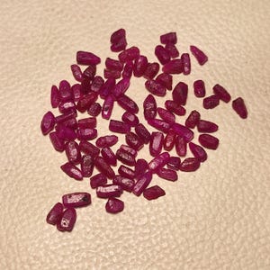 May include: A collection of small, raw ruby gemstones. The stones are a deep, rich red-purple color and have a rough, natural texture. The gemstones are scattered across a neutral-toned surface.