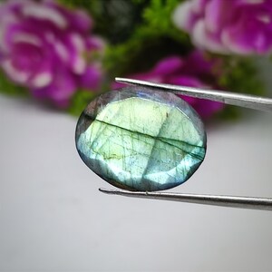 Pode incluir: Uma pedra preciosa de labradorite facetada, em forma oval, segurada por uma pinça. A pedra exibe uma mistura de tons azuis, verdes e amarelos com linhas internas. O fundo apresenta flores rosa desfocadas.