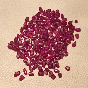 May include: A collection of small, rough-cut ruby gemstones. The gemstones are a deep red colour, with variations in tone and shape. The stones are scattered across a light-coloured surface.
