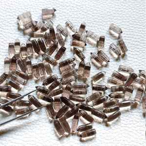 May include: A collection of smoky quartz crystal points, also known as smoky topaz, in various sizes and shapes. The crystals are a dark brown color and have a translucent appearance. The crystals are scattered on a white surface.