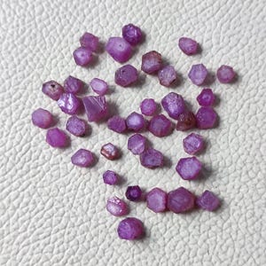Könnte beinhalten: Eine Sammlung roher, ungeschliffener Rubin-Edelsteine. Die Edelsteine haben eine tiefrosa-rote Farbe mit einer rauen, kristallinen Textur. Die Rubine variieren in Größe und Form und sind auf einer weißen Oberfläche verstreut.
