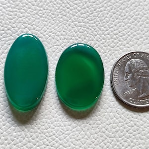 Può includere: Due cabochon di agata verde di forma ovale, uno più grande dell'altro, accanto a un quarto di dollaro americano per il confronto delle dimensioni.