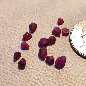 May include: A collection of small, red ruby gemstones, some with a slightly darker hue, scattered on a beige surface. A US quarter is included for scale.