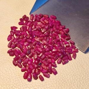 May include: A scattering of small, rough-cut ruby gemstones in shades of red and pink. The gemstones are displayed on a light surface, with a metal tool partially visible. The rubies have a natural, unpolished appearance, showcasing their raw beauty.