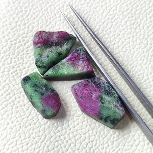 May include: Four rough-cut ruby and zoisite gemstones with a mix of deep pink and green hues. The gemstones are irregularly shaped and have a natural, unpolished appearance. A pair of silver tweezers is positioned near the stones.